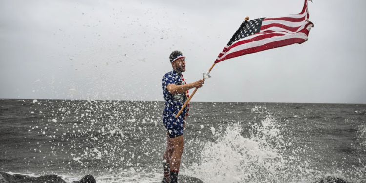 El youtuber Mark Peyton sostiene una bandera estadounidense mientras posa para su hermano Matt Peyton en la costa antes de la llegada del huracán Helene a Alligator Point, Florida, el 26 de septiembre de 2024.